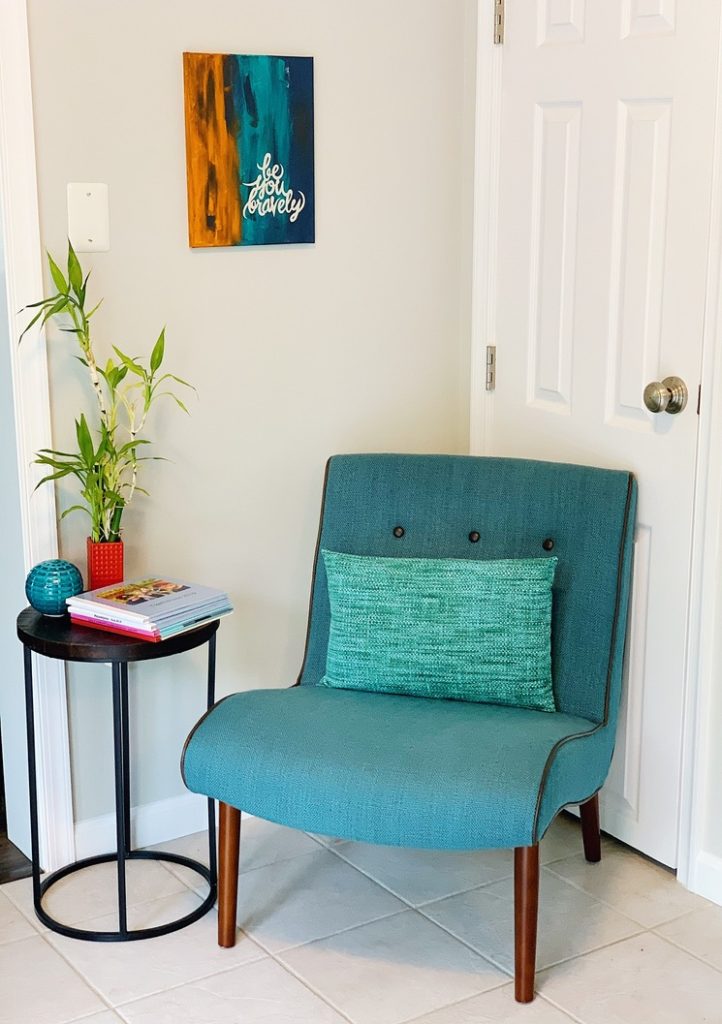 Teal chair with a small pillow. Next tothe chair is a small circular table with a plant on it and a stack of books. Above the chair is a painting that says "be bravely you"