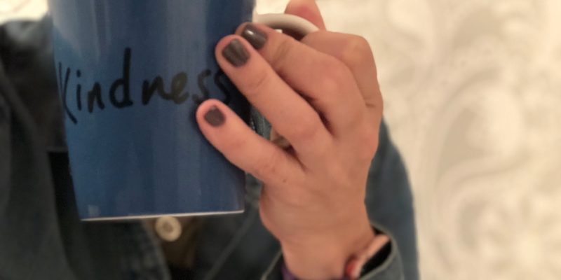 woman holding a coffee mug up to her lips and the mug says kindness her nails are painted a dark gray