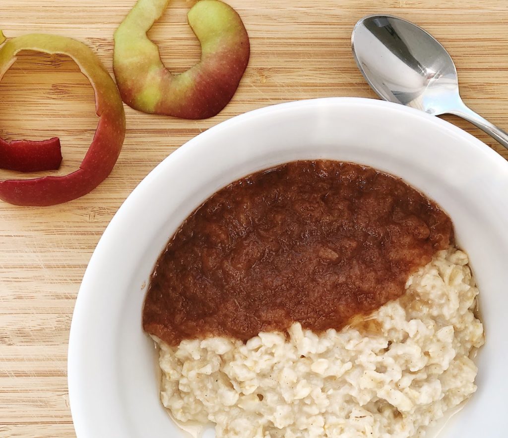 Applesauce in a white bowl with oatmeal and a spoon is in the corner with apple slices
