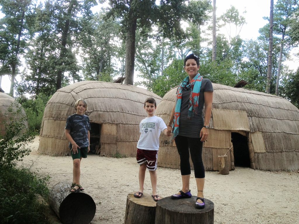 Gretchen holding the hand of her younger kid with her older one slightly off to the side. They are standing in front of Native American Powhatan longhouses.