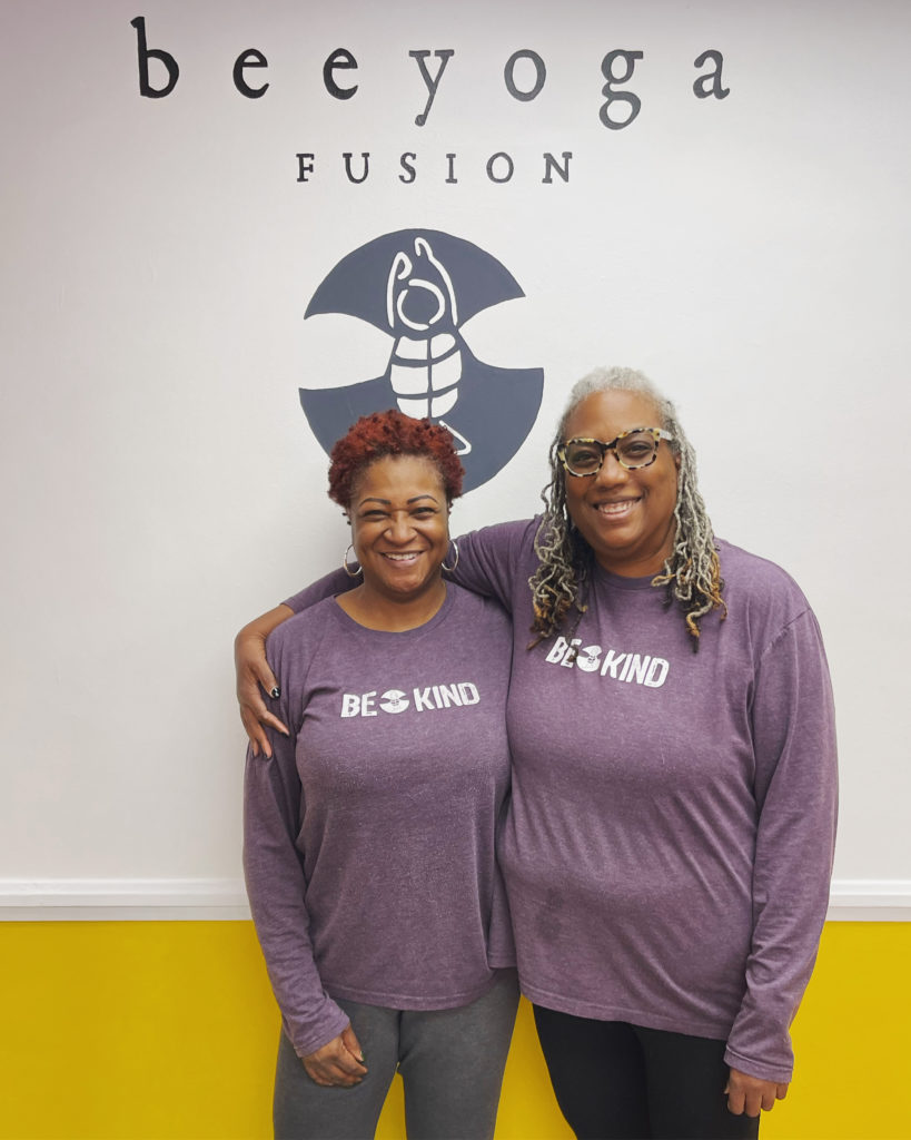 two women wearing the same shirt that says "Be Kind" standing in front of the logo for Bee Yoga Fusion